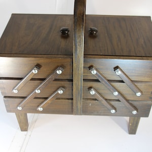 Puede incluir: Una caja de costura de madera vintage con acabado marr&oacute;n oscuro. La caja tiene una tapa con bisagras con dos perillas negras y un asa. La parte delantera presenta bandejas extra&iacute;bles con detalles met&aacute;licos decorativos. La caja mide unos 30 cm de altura.