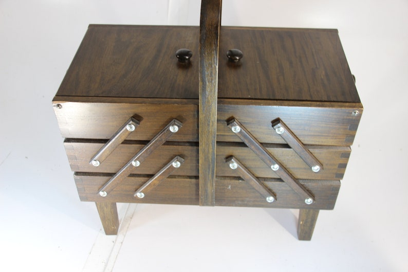 Puede incluir: Una caja de costura de madera vintage con acabado marr&oacute;n oscuro. La caja tiene una tapa con bisagras con dos perillas negras y un asa. La parte delantera presenta bandejas extra&iacute;bles con detalles met&aacute;licos decorativos. La caja mide unos 30 cm de altura.