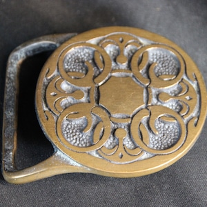 May include: A vintage brass belt buckle with an ornate, circular design. The buckle features a central rectangular shape surrounded by swirling patterns and a textured background. The buckle's frame is a lighter brass color, and the overall design is detailed.