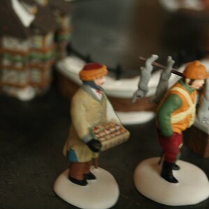 May include: Miniature figurines of two people in winter attire. One figure carries a basket of goods, the other carries a pole with items over their shoulder. The figures are on white bases, with a blurred background of buildings.