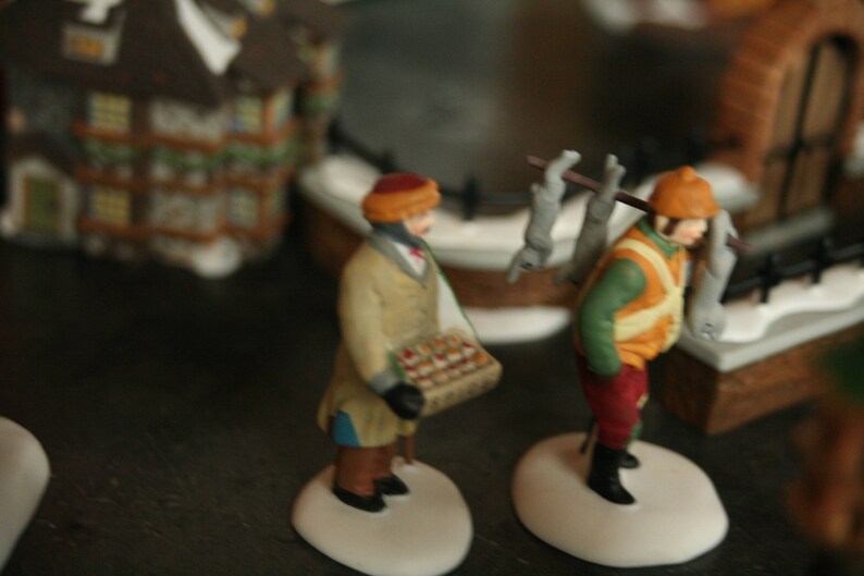 May include: Miniature figurines of two people in winter attire. One figure carries a basket of goods, the other carries a pole with items over their shoulder. The figures are on white bases, with a blurred background of buildings.