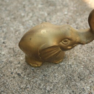 May include: A small, brass elephant ornament with its trunk raised. The elephant has detailed features and a slightly aged appearance. The ornament is resting on a textured, grey surface.