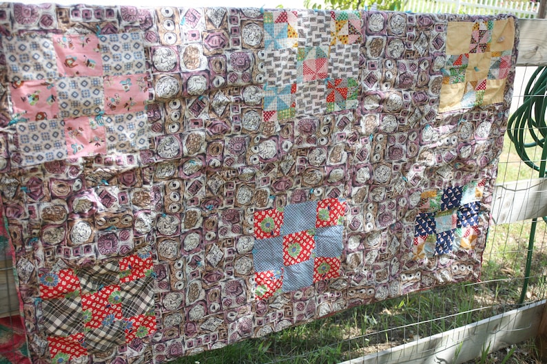 May include: A handmade quilt with a patchwork design, featuring squares of various patterns and colors. The quilt is primarily brown with accents of pink, red, blue, and yellow. It is hanging outdoors, showcasing its intricate details and craftsmanship.
