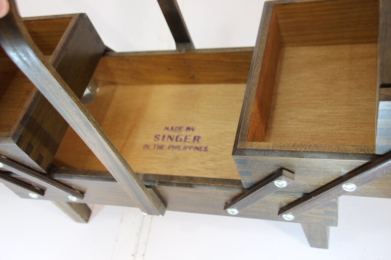 Puede incluir: Una caja de costura de madera vintage con m&uacute;ltiples compartimentos y una tapa con bisagras. La caja es de madera marr&oacute;n oscuro y presenta el texto "MADE BY SINGER IN THE PHILIPPINES" en letras moradas. La caja est&aacute; abierta, revelando los compartimentos interiores.