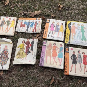 May include: A collection of vintage McCall's sewing patterns. The patterns feature illustrations of dresses and other garments in various colors. The covers are in various colors, including orange, yellow, pink, and blue. The patterns are arranged on a bed of dry grass.