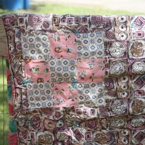 May include: A patchwork quilt with a variety of patterned squares in shades of pink, brown, and cream. The quilt features geometric and floral designs, with a central pink cross pattern. The quilt is draped over a wooden fence.