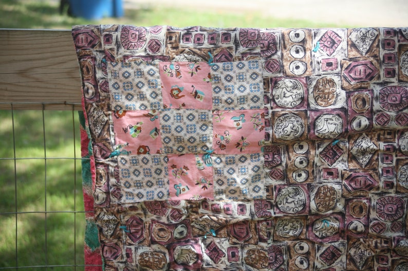 May include: A patchwork quilt with a variety of patterned squares in shades of pink, brown, and cream. The quilt features geometric and floral designs, with a central pink cross pattern. The quilt is draped over a wooden fence.