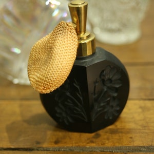 May include: A vintage black perfume bottle with a gold-colored atomizer and a woven gold-colored puff. The bottle has a floral design and sits on a wooden surface. Decorative and elegant.
