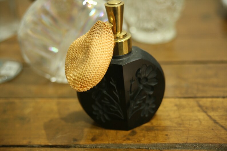 May include: A vintage black perfume bottle with a gold-colored atomizer and a woven gold-colored puff. The bottle has a floral design and sits on a wooden surface. Decorative and elegant.
