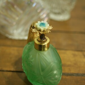 May include: A vintage green perfume bottle with a floral design. The bottle has a gold-colored metal top and a decorative flower-shaped stopper with blue accents. The bottle is likely made of glass.