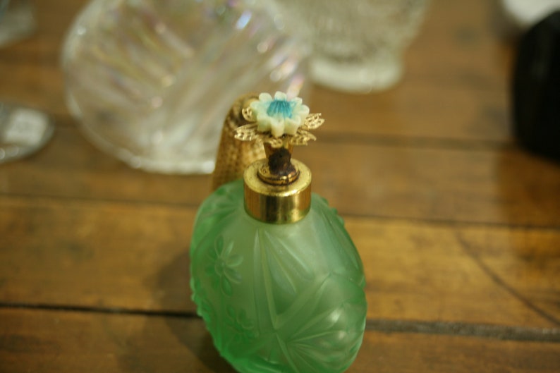 May include: A vintage green perfume bottle with a floral design. The bottle has a gold-colored metal top and a decorative flower-shaped stopper with blue accents. The bottle is likely made of glass.