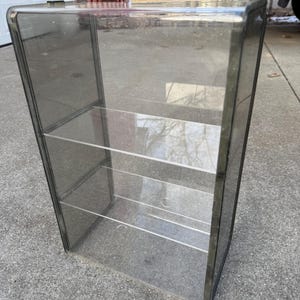 May include: A clear, rectangular display case with three shelves. The case has a dark gray frame and is made of transparent material, allowing full visibility of the contents. The case is designed to stand upright and is suitable for showcasing collectibles or merchandise.