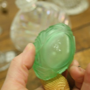 May include: A light green, glass perfume bottle stopper with a textured, leaf-like design. The stopper is held up against a blurred background of other glass objects. The base of the stopper is a woven, tan material.
