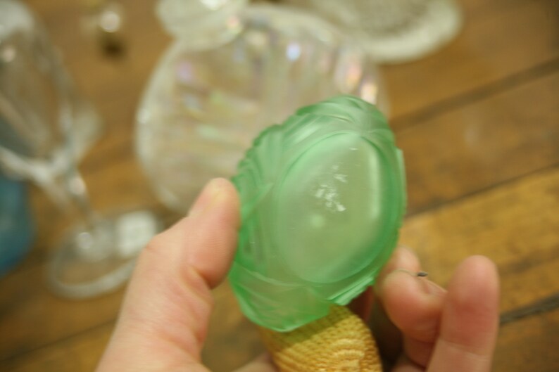 May include: A light green, glass perfume bottle stopper with a textured, leaf-like design. The stopper is held up against a blurred background of other glass objects. The base of the stopper is a woven, tan material.