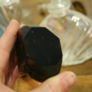 May include: A black, faceted, geometric object is held in a hand. The object has multiple flat sides and a dark, matte finish. Other glass bottles are visible in the background.