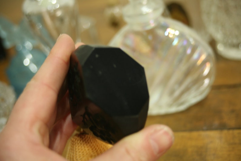 May include: A black, faceted, geometric object is held in a hand. The object has multiple flat sides and a dark, matte finish. Other glass bottles are visible in the background.