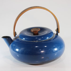 May include: A blue enamel teapot with a wooden handle and lid knob. The teapot has a rounded shape and a spout for pouring. The handle is made of light brown wood and is attached to the pot with metal brackets. The lid is also blue with a small wooden knob.