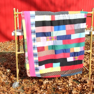 May include: A colorful patchwork quilt displayed on a vintage yellow rack outdoors. The quilt features a variety of fabric patterns and colors, including red, black, blue, and pink. The quilt is rectangular and appears to be handmade.