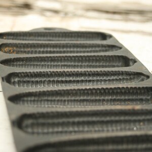 May include: A cast iron cornbread pan with seven corn cob-shaped molds. The pan is dark gray with a textured surface, showing signs of use. The design allows for individual cornbread servings, perfect for baking.