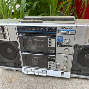 May include: Silver Emerson boombox with black speaker grilles, cassette decks, and radio dial. The boombox has a handle, buttons, and dials for volume and tuning. The radio dial displays FM and AM frequencies.