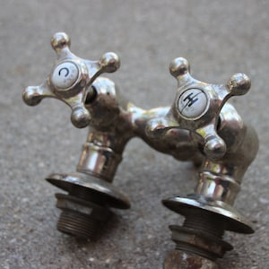 Könnte beinhalten: Nahaufnahme eines antiken, silberfarbenen Wasserhahns mit kreuzförmigen Griffen. Die Griffe haben weiße, kreisförmige Einsätze mit schwarzen Markierungen. Der Wasserhahn weist Alterserscheinungen und Abnutzungserscheinungen auf, mit sichtbarem Rost und Verfärbungen.