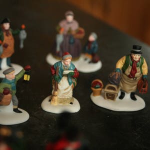 May include: A collection of miniature Christmas village figurines. The figurines depict various characters, including a man with a lantern, a woman with a basket, and a man with a top hat. Each figurine is on a white base.