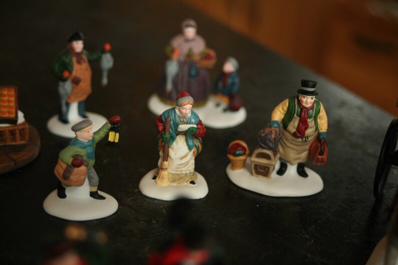 May include: A collection of miniature Christmas village figurines. The figurines depict various characters, including a man with a lantern, a woman with a basket, and a man with a top hat. Each figurine is on a white base.