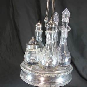 Pode incluir: Um conjunto de condimentos prateado com garrafas e potes de vidro transparente. O conjunto inclui várias garrafas com rolhas e um pote pequeno, todos apoiados numa base prateada decorativa em camadas. As peças de vidro têm desenhos gravados.