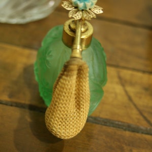 May include: A vintage perfume bottle with a green glass body and a gold-colored metal atomizer. The atomizer is topped with a flower design in gold, white, and blue. The bottle has a woven beige bulb. The bottle is on a wooden surface.