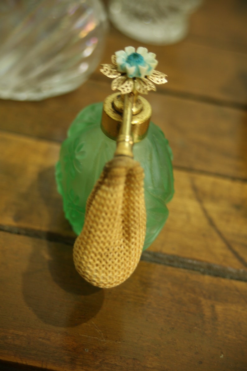 May include: A vintage perfume bottle with a green glass body and a gold-colored metal atomizer. The atomizer is topped with a flower design in gold, white, and blue. The bottle has a woven beige bulb. The bottle is on a wooden surface.
