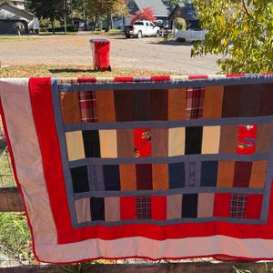 May include: A handmade quilt with a patchwork design, featuring rectangular blocks in various colors and patterns. The quilt has a red border and is displayed outdoors on a wooden fence. The quilt's dimensions are not visible.