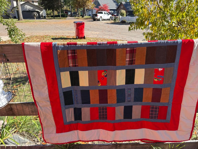 May include: A handmade quilt with a patchwork design, featuring rectangular blocks in various colors and patterns. The quilt has a red border and is displayed outdoors on a wooden fence. The quilt's dimensions are not visible.