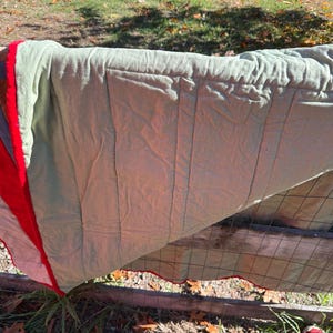 May include: A multi-colored quilt draped over a wooden fence. The quilt features red, white, blue, and green sections. The quilt has a scalloped red edge and is made of a soft, textured fabric. The quilt is outdoors in natural light.