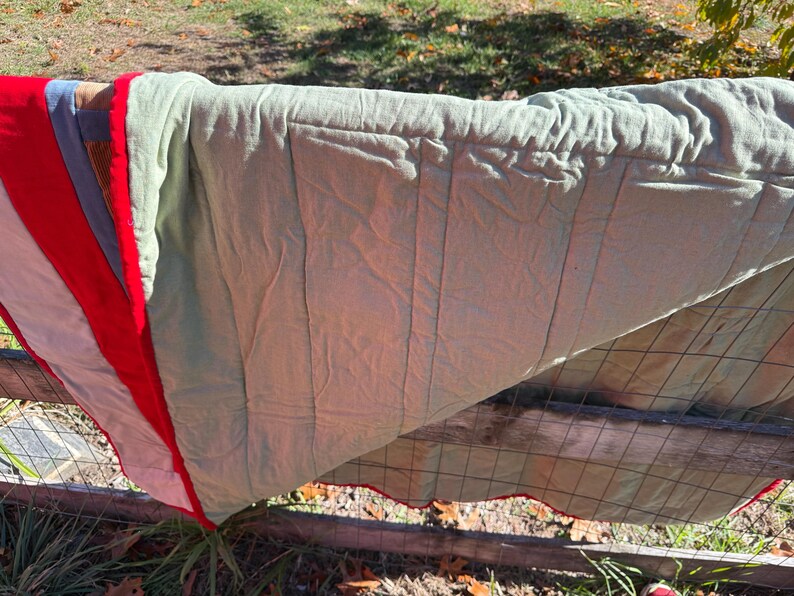 May include: A multi-colored quilt draped over a wooden fence. The quilt features red, white, blue, and green sections. The quilt has a scalloped red edge and is made of a soft, textured fabric. The quilt is outdoors in natural light.