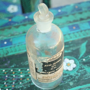 May include: A clear glass bottle with a glass stopper. The bottle has a label that reads "Acid Nitric" and "Poison". The bottle is 454 grams in size. The bottle is sitting on a teal and blue surface.