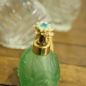May include: A vintage green glass perfume bottle with a gold-colored cap and a decorative flower-shaped stopper. The bottle has an embossed floral design and sits on a wooden surface. Other glass objects are in the background.