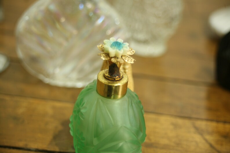 May include: A vintage green glass perfume bottle with a gold-colored cap and a decorative flower-shaped stopper. The bottle has an embossed floral design and sits on a wooden surface. Other glass objects are in the background.