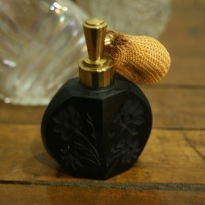 May include: Black perfume bottle with a gold-colored atomizer and a woven tan bulb. The bottle has a floral design carved into the surface. The bottle is sitting on a wooden surface.