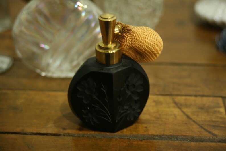 May include: Black perfume bottle with a gold-colored atomizer and a woven tan bulb. The bottle has a floral design carved into the surface. The bottle is sitting on a wooden surface.