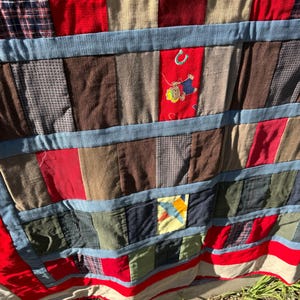 May include: A patchwork quilt with a variety of fabrics in red, blue, brown, and beige. The quilt features a blue border and an embroidered patch with a cartoon figure. The quilt is made of various textures and patterns.