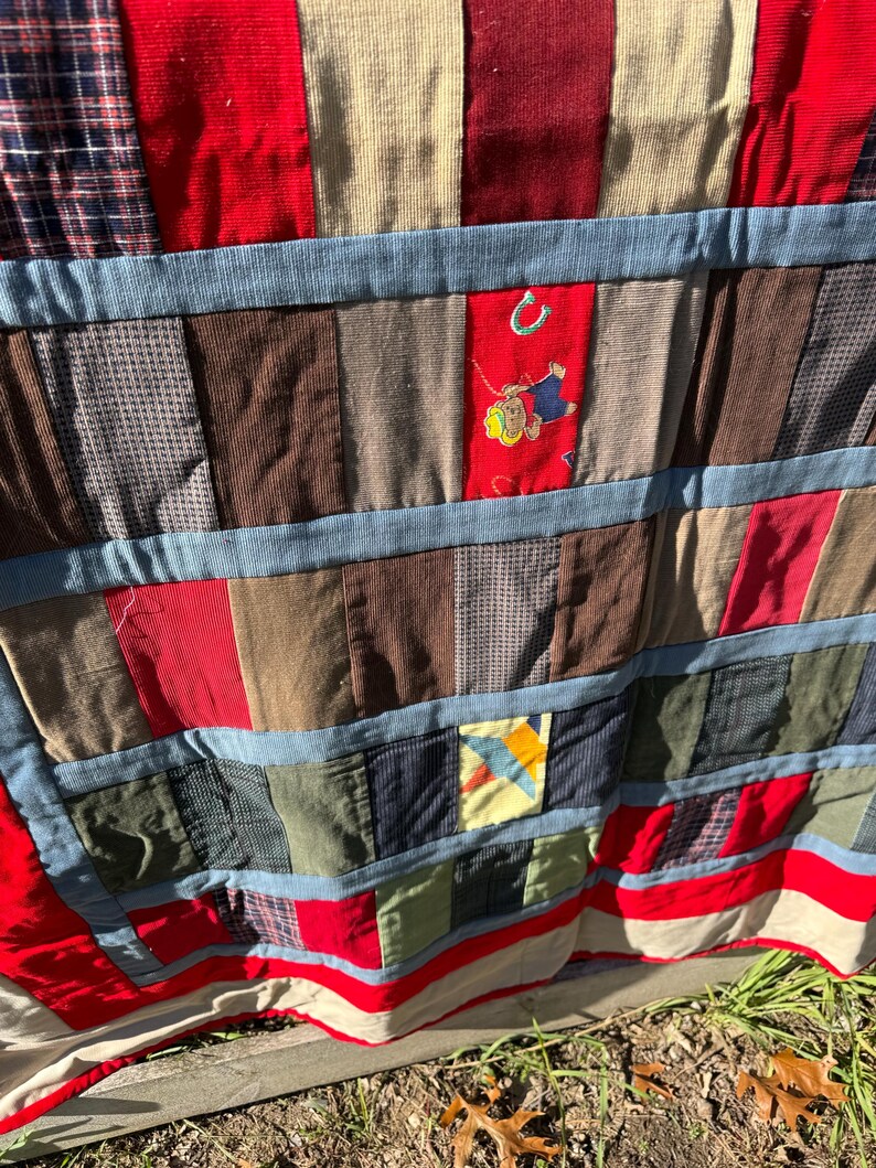 May include: A patchwork quilt with a variety of fabrics in red, blue, brown, and beige. The quilt features a blue border and an embroidered patch with a cartoon figure. The quilt is made of various textures and patterns.