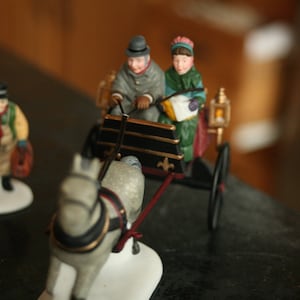 May include: A miniature Christmas scene featuring a horse-drawn carriage with two figures, and another figure standing nearby. The carriage is black and gold, and the figures are dressed in winter attire. The horse is gray and white.