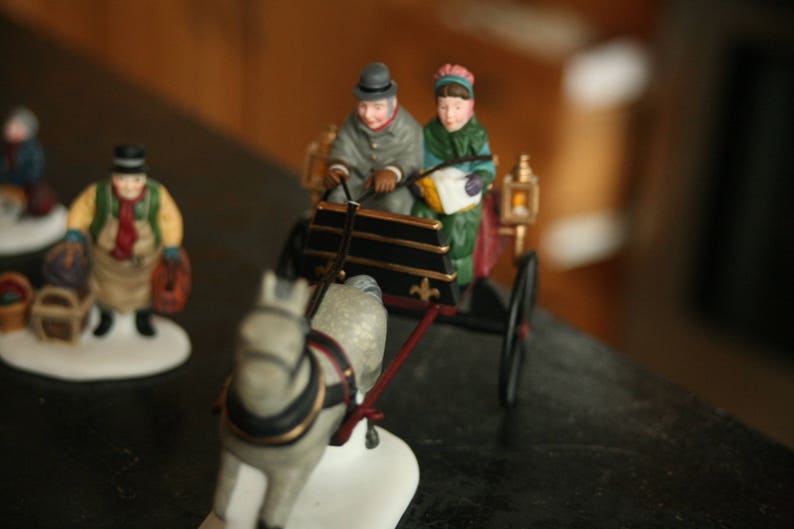 May include: A miniature Christmas scene featuring a horse-drawn carriage with two figures, and another figure standing nearby. The carriage is black and gold, and the figures are dressed in winter attire. The horse is gray and white.