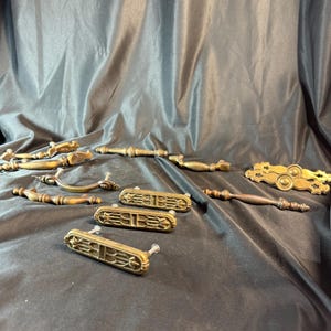 Könnte beinhalten: Eine Sammlung antiker Messing-Schrankbeschläge, darunter Schubladenzüge und Türknauf. Die Beschläge weisen komplizierte Designs und eine abgenutzte, gealterte Oberfläche auf.