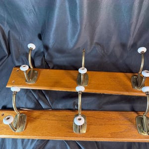 May include: Two sets of vintage coat racks. Each rack has a rectangular wooden base with three brass hooks and white ceramic knobs. The wood has a warm, natural finish. The hooks are mounted on a dark background.