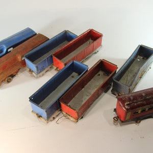May include: Collection of vintage toy train cars in various colors, including blue, red, and maroon. The train cars are made of metal and show signs of wear, suggesting they are antique or well-loved. The collection includes several open-top cargo cars and a passenger car.