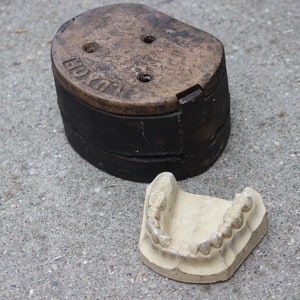 Könnte beinhalten: Ein antikes Dentalformenset, mit einer dunklen, verwitterten Metallbasis, auf deren Deckel das Wort "LUXOR" eingeprägt ist. Ein Gips-Zahnabdruck liegt neben der Basis und zeigt die Form der oberen Zähne.