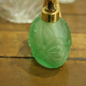 May include: A green glass perfume bottle with a gold-colored metal top. The bottle has a frosted finish and a detailed floral design. The bottle is sitting on a wooden surface.