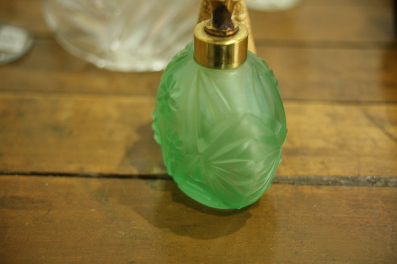 May include: A green glass perfume bottle with a gold-colored metal top. The bottle has a frosted finish and a detailed floral design. The bottle is sitting on a wooden surface.