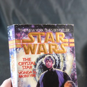 May include: A paperback book titled "Star Wars: The Crystal Star" by Vonda N. McIntyre. The cover features a space scene with a portrait of a man. The title is in gold lettering, and the book is a New York Times bestseller.